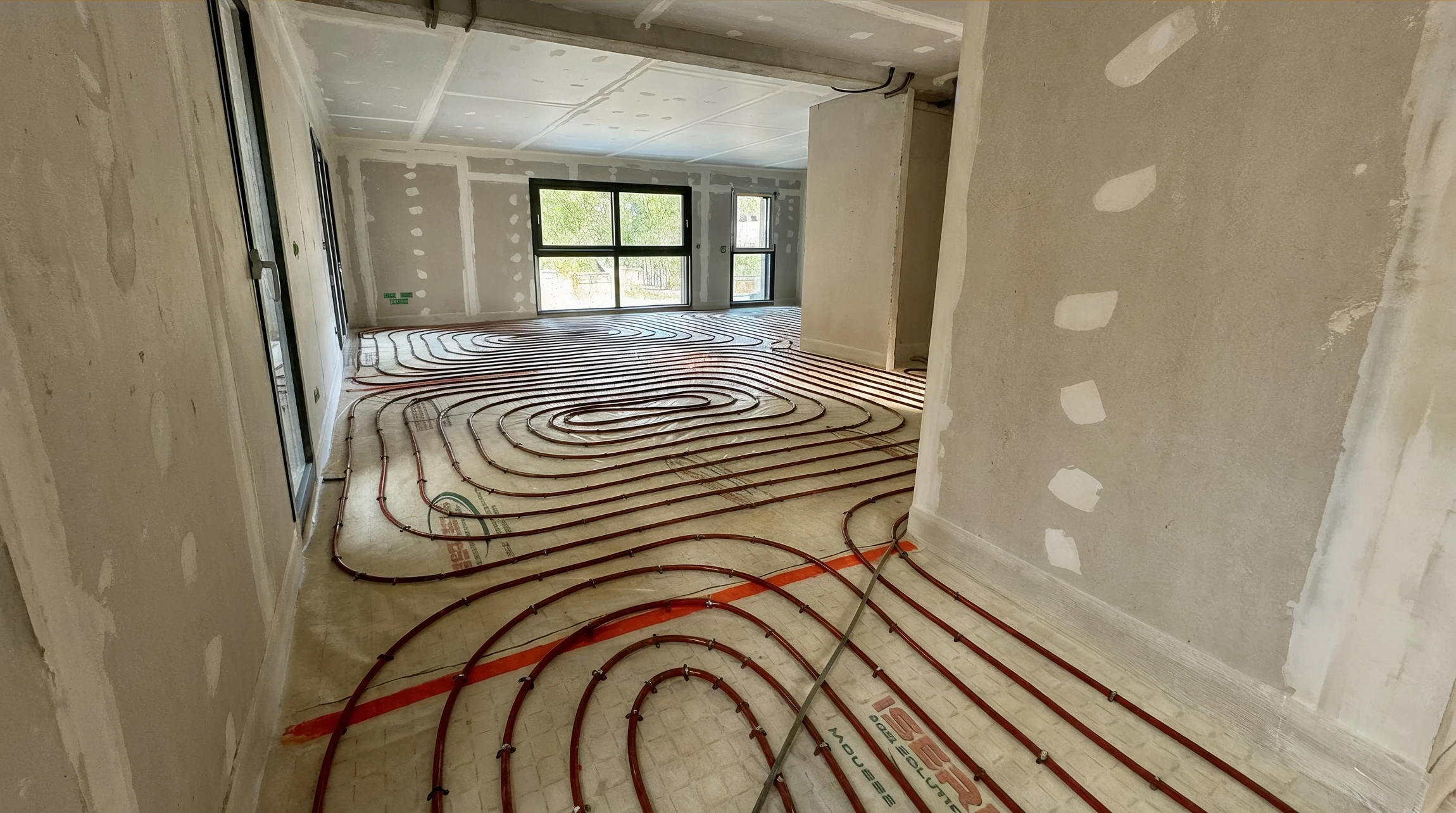 Intérieur résidentiel avec technicien en combinaison rouge et bleue installant un radiateur blanc à ailettes sur mur gris